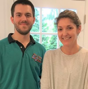 A man in a teal polo shirt stands next to a woman in a light gray top, both smiling, indoors in front of a window with green trees visible outside.