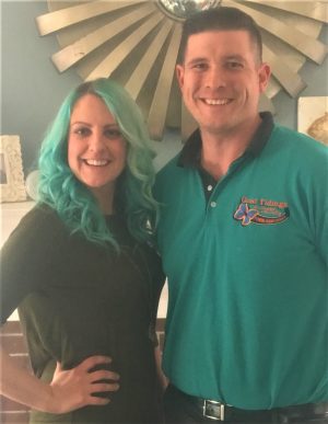 A woman with long teal hair and a man in a teal shirt stand side by side, smiling indoors in front of a decorative mirror.