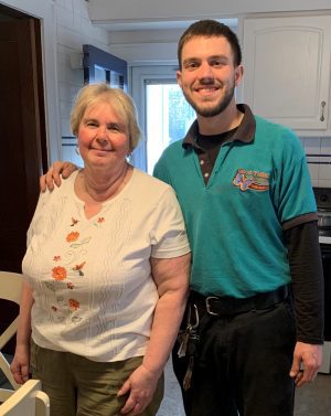 An older woman and a younger man stand side by side in a kitchen, smiling at the camera. The man has his arm around the woman's shoulder.