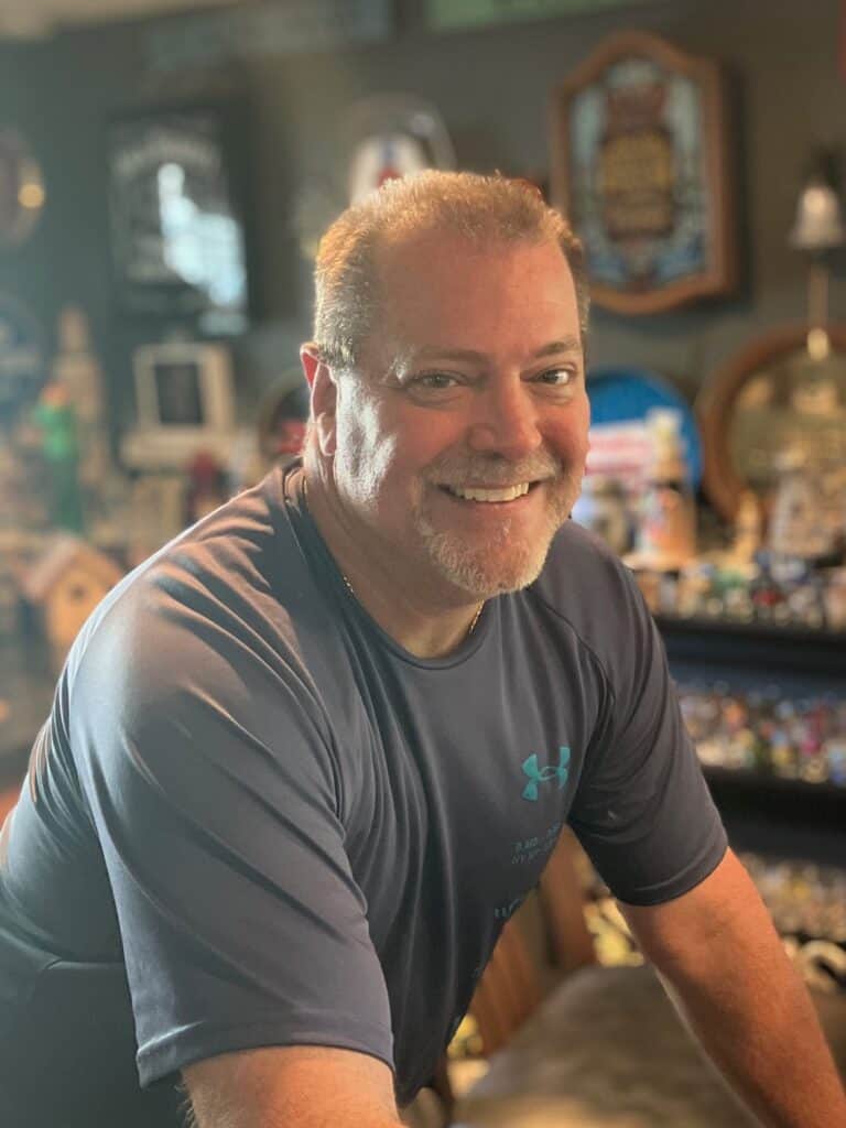 A middle-aged man with short hair and a beard smiles while leaning forward in a room filled with various collectibles and decorations.