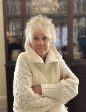 Older woman with white hair and a serious expression stands indoors with her arms crossed, wearing a white knit sweater. China cabinet with dishes is visible in the background.