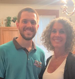Two people standing indoors, smiling at the camera. The man is wearing a teal shirt, and the woman has curly hair and is wearing a white top with a black cardigan.