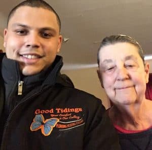 Two people pose for a photo indoors. The person on the left wears a black jacket with "Good Tidings" and a butterfly logo, while the person on the right wears a red-trimmed top.