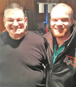 Two men are standing side by side indoors, both smiling at the camera. One is wearing a black shirt, and the other is wearing a dark jacket with a logo and green shirt.