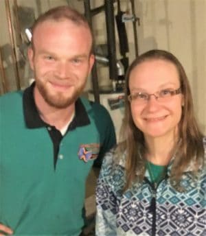 Two people stand indoors, smiling at the camera. The man wears a green shirt with a logo, and the woman wears glasses and a patterned sweater. Shelves and equipment are visible in the background.