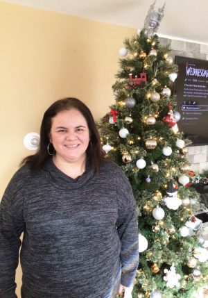 A woman in a gray sweater stands smiling beside a decorated Christmas tree indoors.