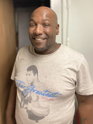 A man smiles at the camera while wearing a beige t-shirt featuring an illustration of a boxer and the text "The Greatest of All Time.