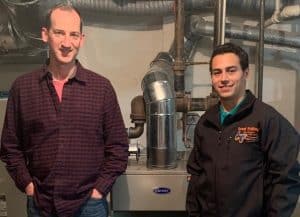Two men stand in front of a large HVAC unit in a basement or utility room, with pipes and ducts visible in the background.