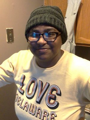 A person wearing glasses, a dark knit hat, and a beige sweatshirt with the word "LOVE" and "DELAWARE" stands indoors, smiling at the camera.