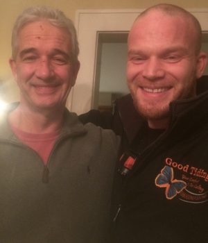 Two men are standing indoors, smiling at the camera, with one man’s arm around the other’s shoulder.