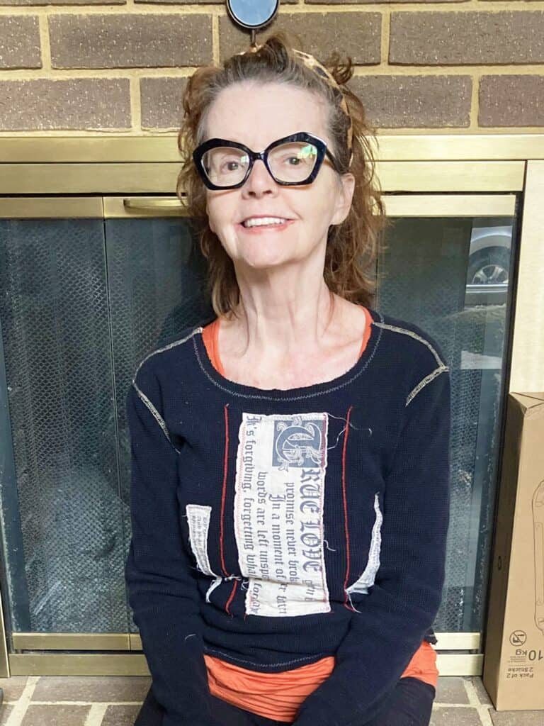 A smiling woman with glasses and a patterned shirt sits in front of a brick fireplace.