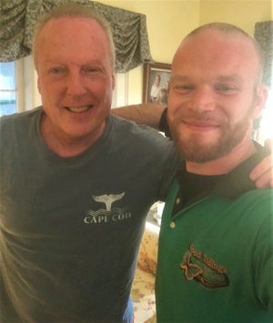 Two men are smiling at the camera indoors; one is wearing a blue Cape Cod shirt, and the other is wearing a green collared shirt.