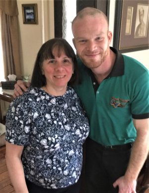 A woman and a man stand close together indoors, smiling at the camera. The woman wears a patterned shirt, and the man wears a green collared shirt.