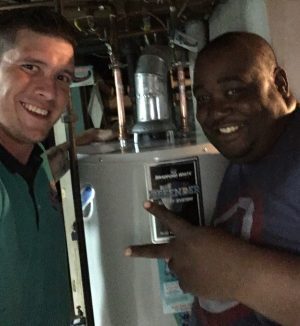 Two men smiling and posing in front of a water heater; one is making a peace sign with his hand.