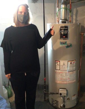 A person wearing a mask stands next to a water heater in a basement, giving a thumbs-up gesture.