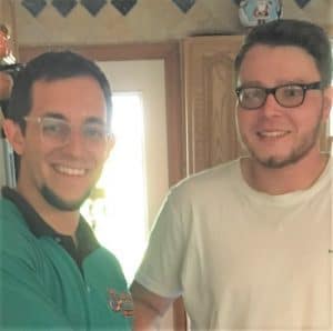 Two men standing indoors, smiling at the camera; one wears glasses and a green shirt, the other wears glasses and a white shirt.
