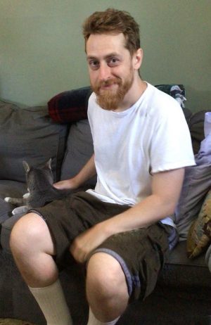 A man with a beard, wearing a white t-shirt and dark shorts, sits on a couch and pets a gray cat beside him.