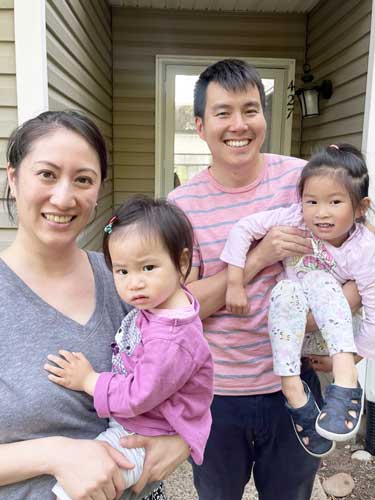 A woman and man stand outside a house, each holding a young child. The adults smile at the camera while the children look at the camera with neutral expressions.