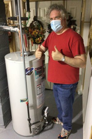 A man wearing a face mask, red shirt, and jeans gives two thumbs up while standing next to a water heater in a basement or utility room.