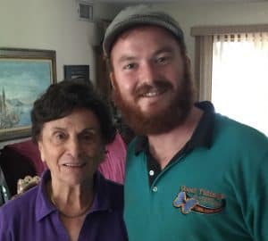 Two people standing indoors, one older woman in a purple shirt and one younger man with a beard and cap wearing a green shirt. They are both smiling at the camera.