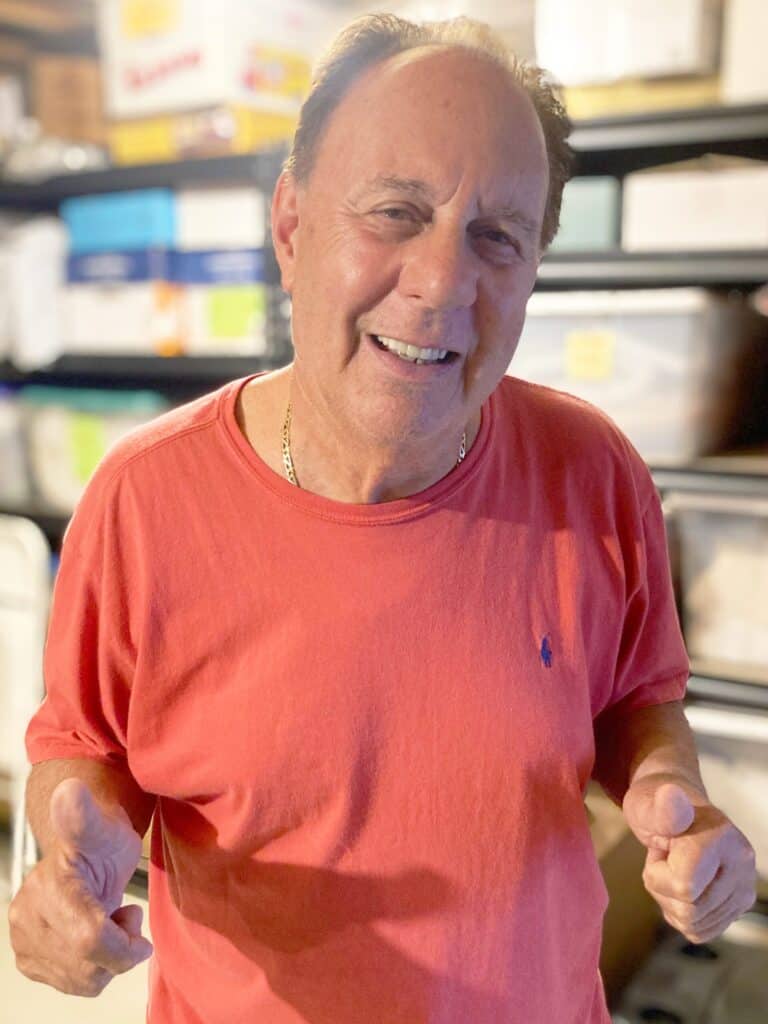 A man in a red shirt stands indoors, smiling at the camera and giving two thumbs up, with shelves of storage boxes in the background.