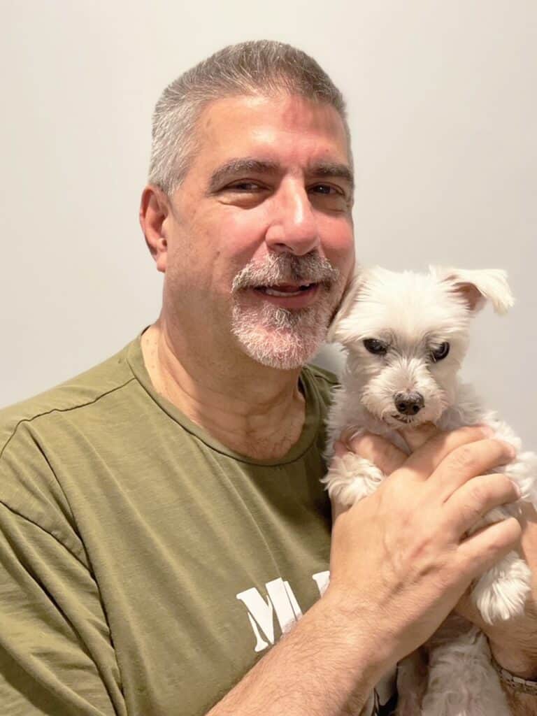 A man with short gray hair and a beard smiles while holding a small white dog close to his chest against a plain background.
