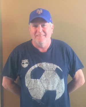 A man wearing a blue baseball cap and a navy t-shirt with a large soccer ball graphic stands in front of a plain beige wall.