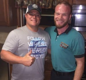 Two men smiling indoors; one wears a gray t-shirt and baseball cap giving a thumbs up, the other wears a teal collared shirt with an embroidered logo.