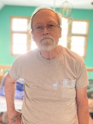 An older man with a white beard and glasses stands indoors in front of a window, wearing a light gray t-shirt. The background is brightly lit with green walls.