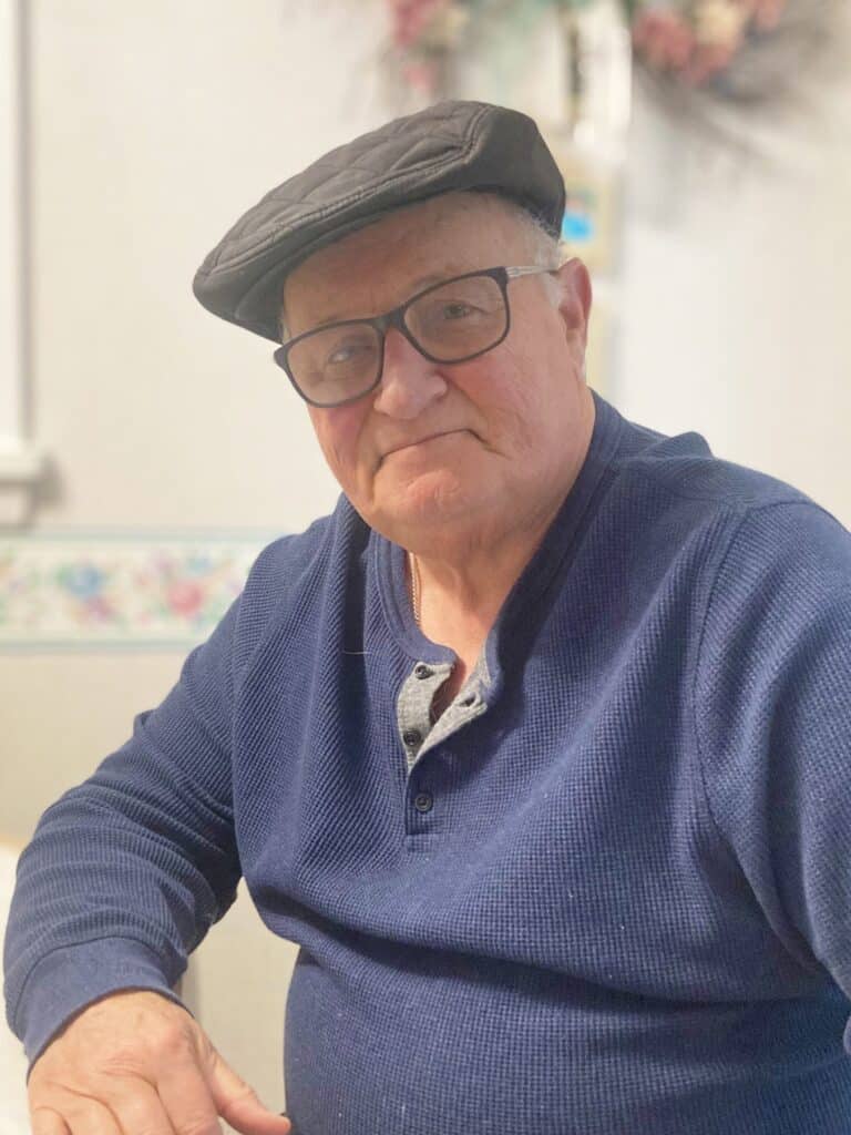 Older man wearing glasses and a flat cap, dressed in a blue shirt, sitting indoors with a neutral expression.