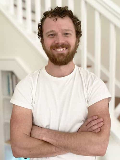 A man with curly hair and a beard, wearing a white t-shirt, stands indoors with his arms crossed and smiles at the camera. A staircase and bookshelves are visible in the background.