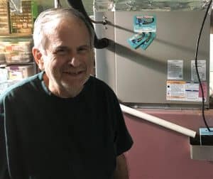 An older man with short gray hair and a beard stands indoors next to an HVAC unit mounted on a wall.