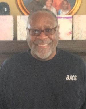 A man wearing glasses and a dark shirt with "BMS" on it stands indoors, smiling at the camera. There are framed pictures in the background.