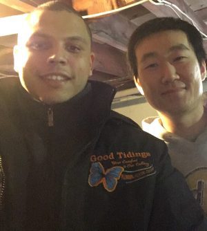 Two men standing indoors, one wearing a dark jacket with a butterfly logo and “Good Tidings” text, and the other wearing a light hoodie, both smiling at the camera.