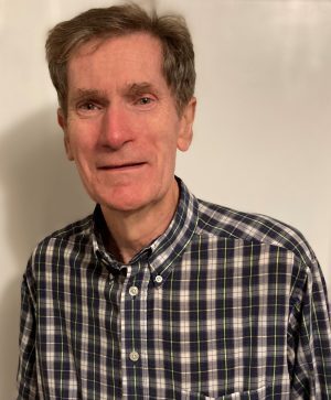 An older man with short brown hair is wearing a plaid button-up shirt and standing in front of a plain light-colored background.