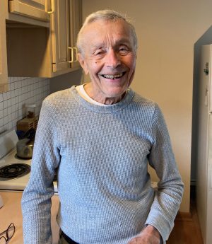 Elderly man with gray hair smiling and standing in a kitchen, wearing a light gray sweater.