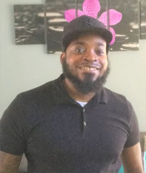 A man wearing a black polo shirt and black cap smiles at the camera, standing indoors with a flower artwork on the wall behind him.