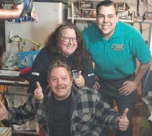 Three people pose indoors, smiling and giving thumbs up. Two stand behind, one kneels in front. Shelves and household items are visible in the background.