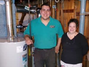 A smiling man in a teal shirt stands next to a woman in a basement beside a water heater and pipes.