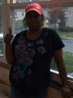 A person wearing a red cap and floral-patterned shirt stands indoors by a window, giving a thumbs-up gesture.