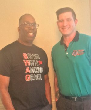 Two men stand side by side indoors, one wearing a black T-shirt reading "Saved With Amazing Grace," and the other in a teal collared shirt.