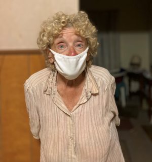 Elderly woman with curly hair wearing a white mask and striped shirt, standing indoors with a neutral expression.