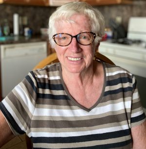 An older woman with short white hair and glasses smiles while sitting in a kitchen, wearing a striped shirt.