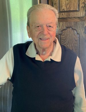 An older man with gray hair and a mustache stands indoors, wearing a light-colored collared shirt under a dark sleeveless sweater, smiling at the camera.