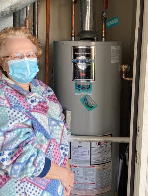An older woman wearing glasses, a mask, and a patterned jacket stands next to a water heater in a utility area.