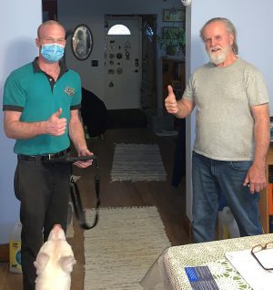 Two men stand indoors giving thumbs up; one man wears a mask and uniform, the other is in casual clothes. A white dog is in the foreground looking up at them.