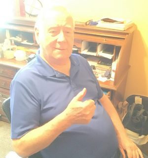 Older man in a blue polo shirt sits at a desk, giving a thumbs-up gesture, with papers and office supplies visible in the background.