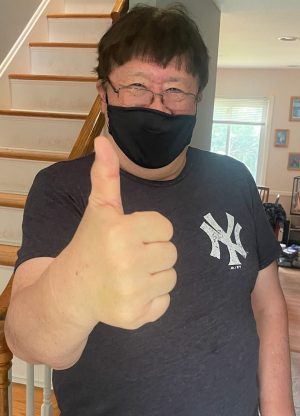 A person wearing a black mask and a New York Yankees t-shirt stands indoors, giving a thumbs-up gesture in front of a staircase.