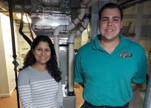 Two people stand in front of HVAC equipment in a basement; the woman is wearing a striped sweater, and the man is wearing a teal polo shirt.