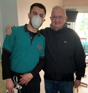 Two men stand indoors; one wears a teal work shirt and face mask, the other wears glasses and a black jacket. The man on the left has keys clipped to his belt.